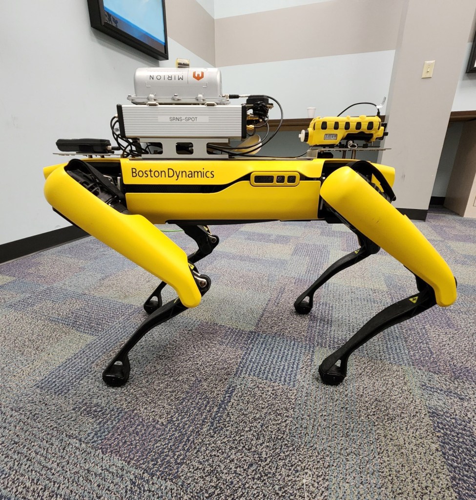 A black and yellow robot dog called Spot walking on geometrical patterned carpet.